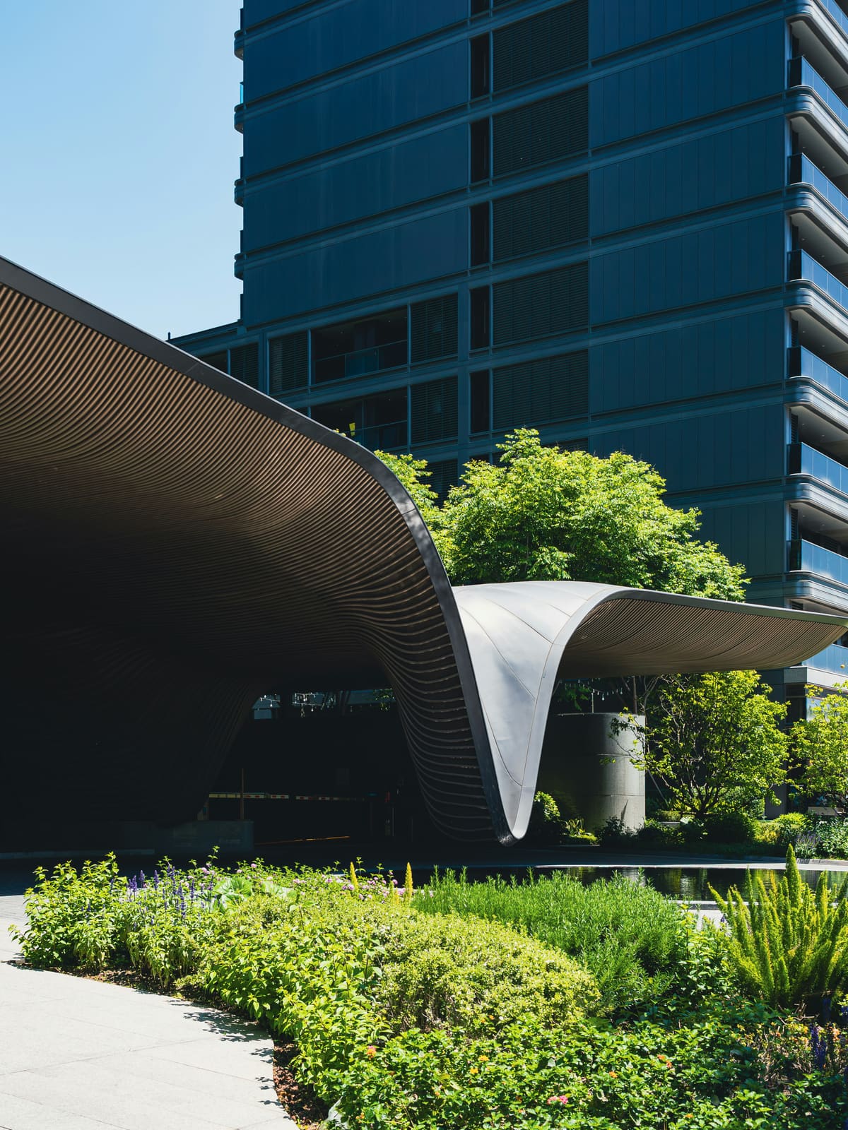 A sweeping curved bronze-toned metal canopy above dense street-level planting, a glass tower rising behind.