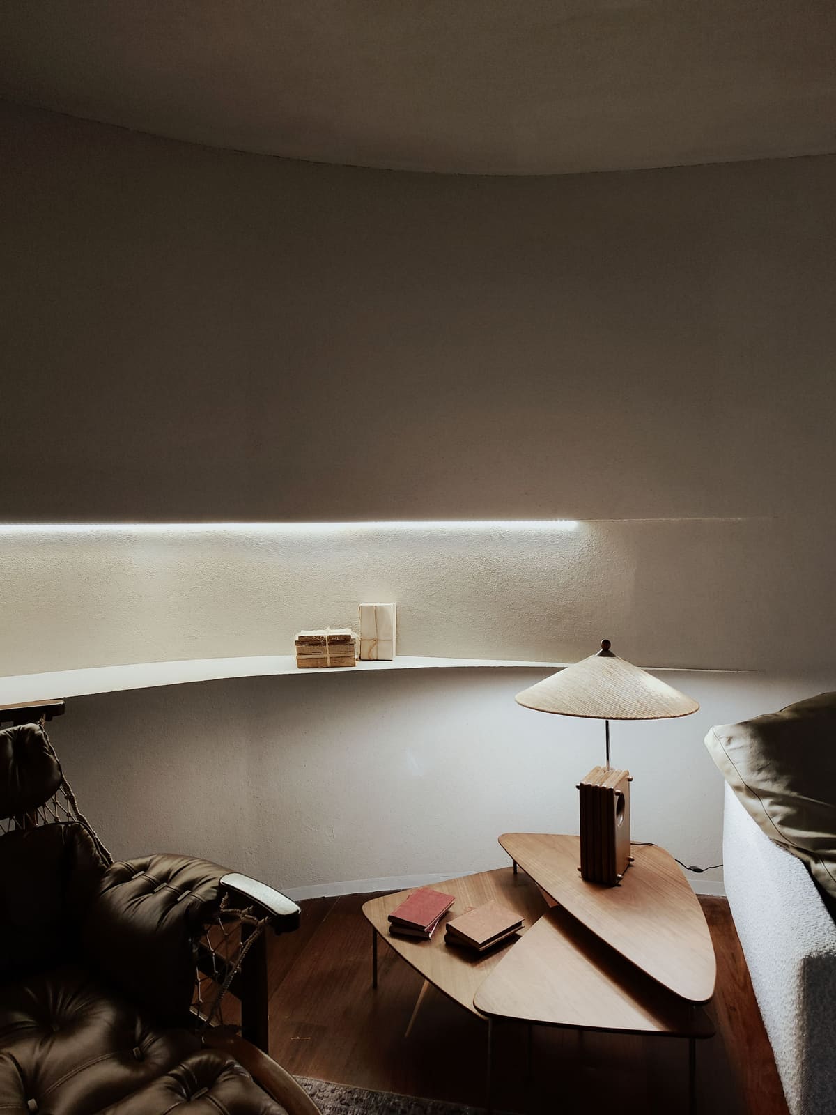 A dark curved room with a warm amber lamp on a low table and a low leather seat.