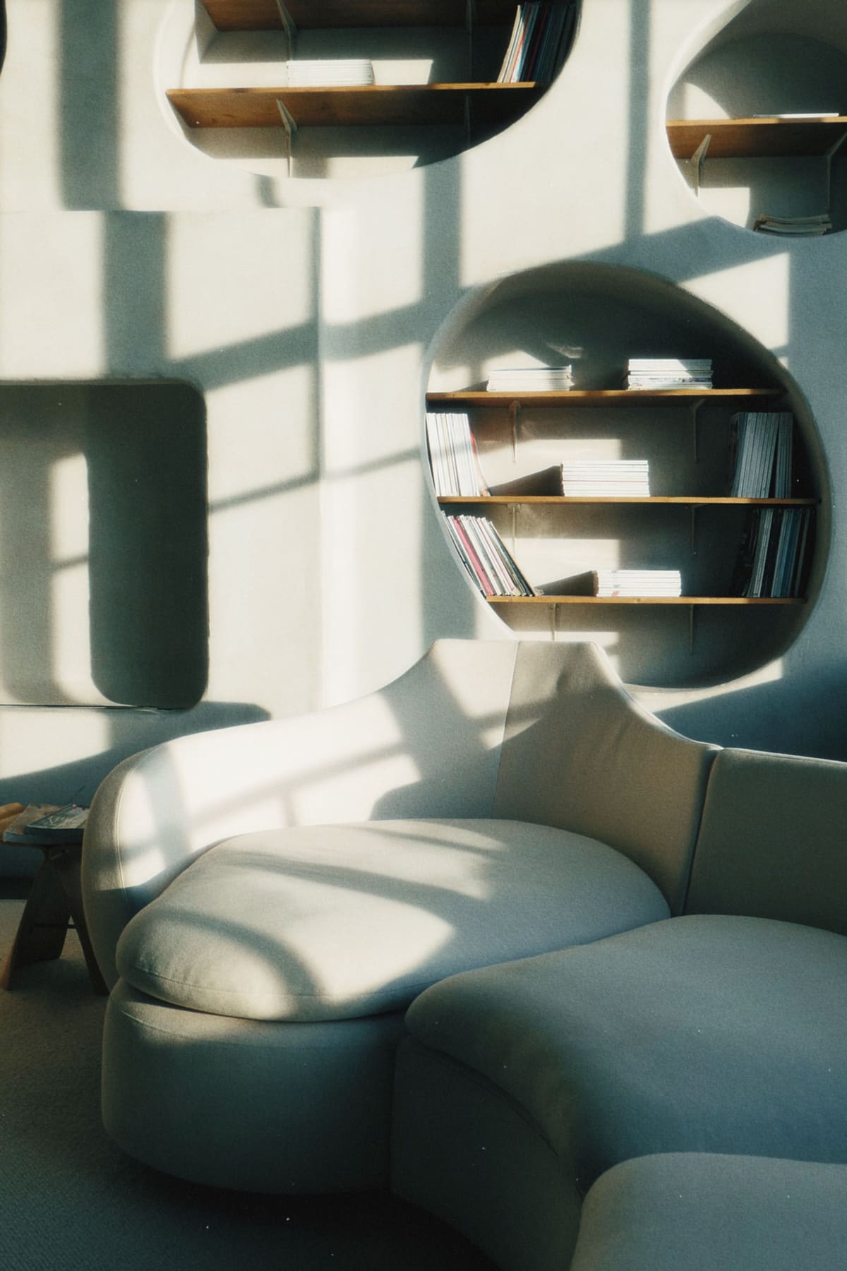 A curved built-in bookshelf and pale sculptural sofa lit by hard window shadow.