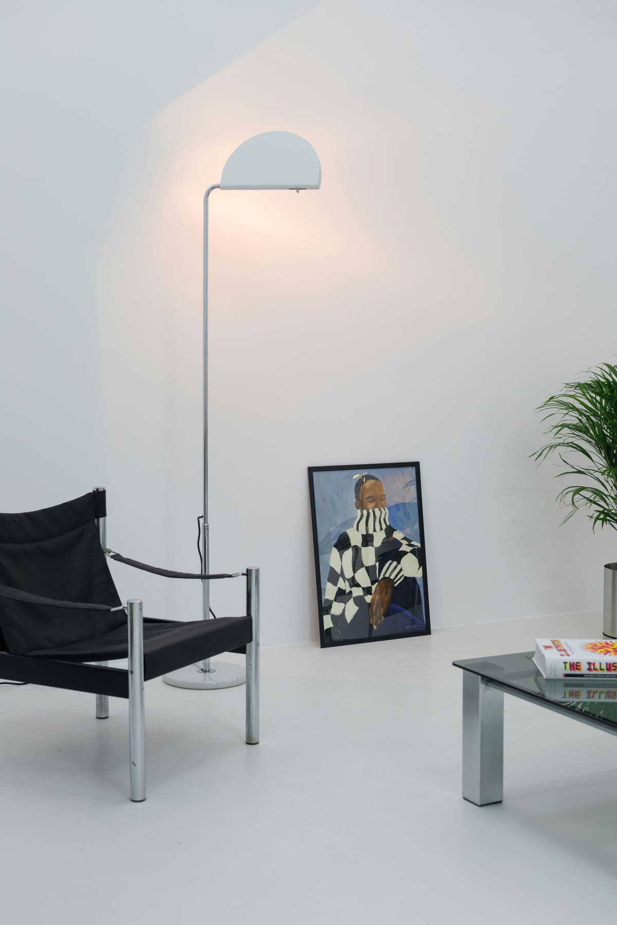 A black sling chair, chrome floor lamp and glass table against a white wall with framed art.
