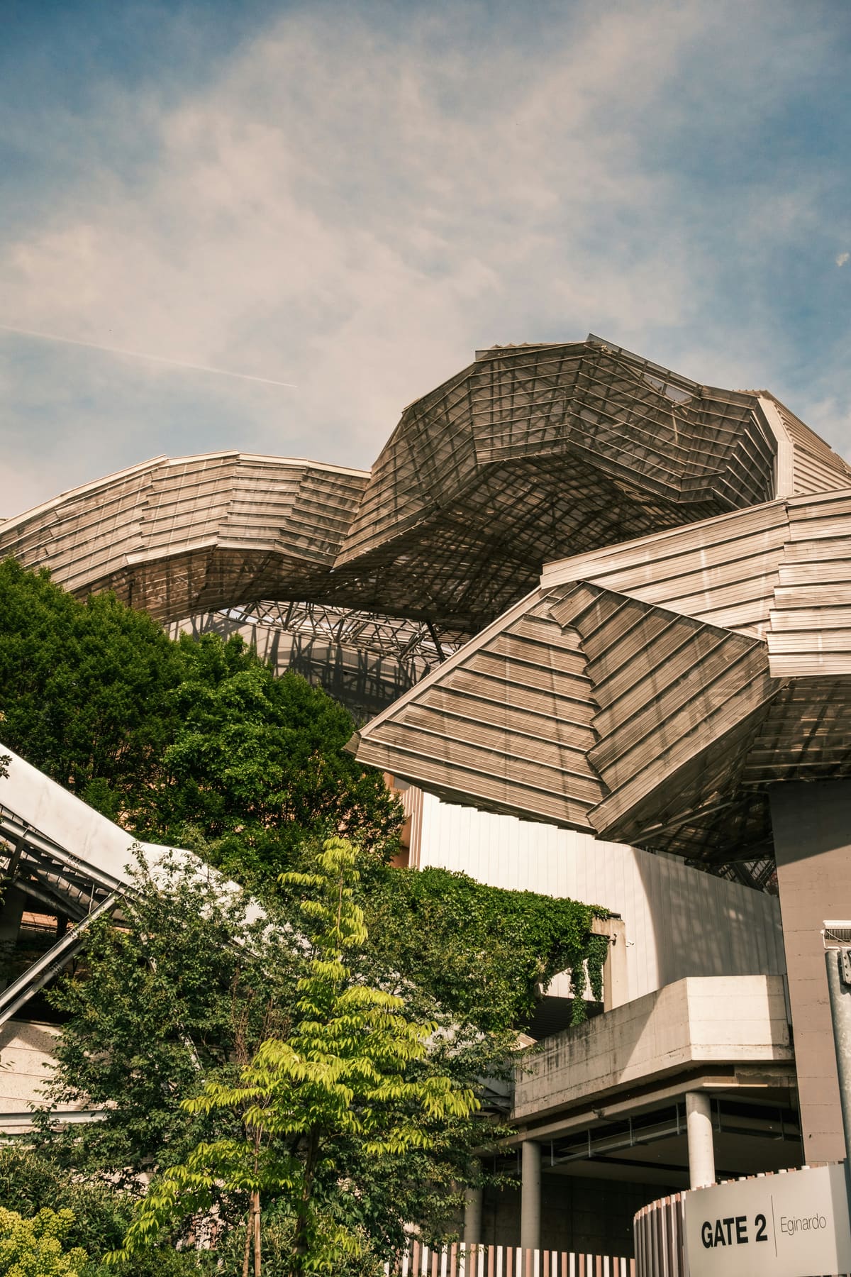 Angular faceted rooflines of a concrete-and-timber structure rising over trees under a soft sky.